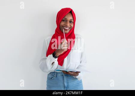 Checklist. Intelligent black muslim lady in hijab taking notes in copybook and thinking, standing on white background Stock Photo