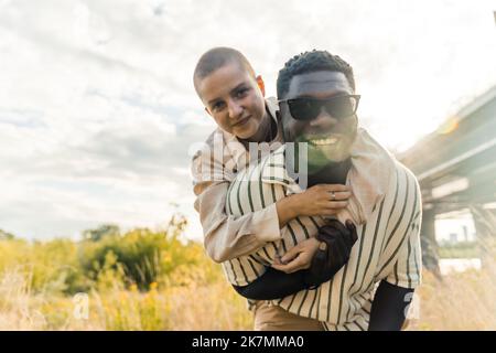 Stunning outdoor portrait of multi-ethnic couple enjoying time outdoors. Handsome tall black man in sunglasses smiling at camera, taking his caucasian bald girlfriend on piggyback. High quality photo Stock Photo