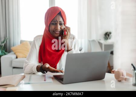 Upset african american woman in hijab looking at laptop screen and talking on phone, having problems with her job Stock Photo
