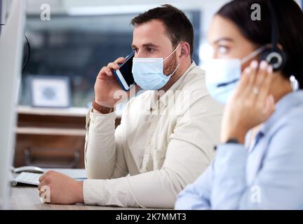 We put safety first in here. a young businessman sitting in the office with his colleague and wearing a face mask while using his cellphone. Stock Photo