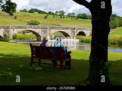 Two women relaxing on bench near the River Wharfe in Burnsall, Wharfedale, North Yorkshire, England UK Stock Photo