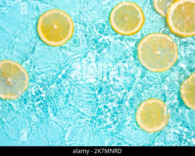 Lemon slices in clean transparent water over blue background with copy space. Water splashing on blue water surface in sunlight. Top view or flatlay. Summer, vacation, healthy eating concept Stock Photo