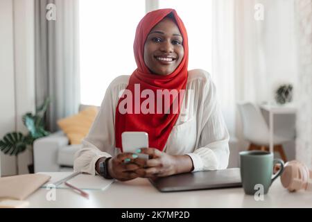 Positive african american muslim woman in hijab using smartphone at home sitting at table and smiling at camera Stock Photo