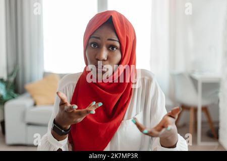 African american muslim woman in hijab talking and looking at camera, having online conversation, sitting at home Stock Photo