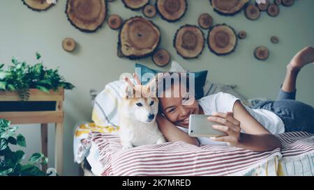 Happy African American girl proud dog owner is taking selfie with cute pet lying on bed in modern apartment using smartphone. Technology, animals and social media concept. Stock Photo