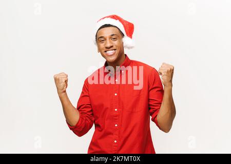 Overjoyed multiracial man in santa hat showing yes gesture on white background. Portrait of cheerful guy raising fists, celebrating victory Stock Photo