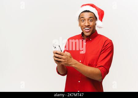 Young handsome happy shocked man in santa hat holds smartphone and looking at the camera with surprised opened mouth, confused cheerful male, isolated on white. New Year advertising concept Stock Photo