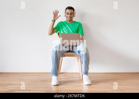 Cheerful arab man in headset having web call via laptop computer, waving hand at webcam, sitting over light wall Stock Photo