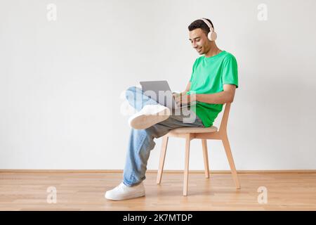 Young male freelancer working with laptop, smiling arab man sitting on chair over light wall, free space, full length Stock Photo