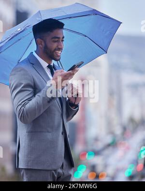 Business happens over the phone. a handsome young businessman standing alone and using his cellphone while holding an umbrella. Stock Photo