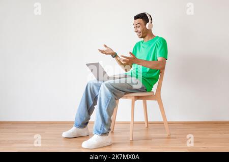 Excited arab man in wireless headphones making video call via laptop while sitting on chair over light wall, copy space Stock Photo
