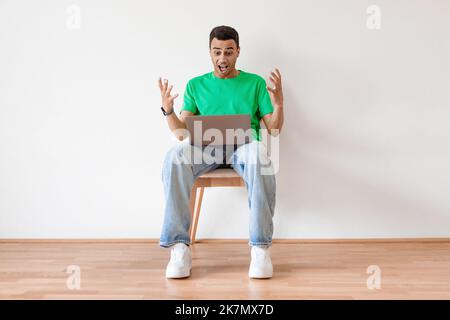Online offer. Emotional arab man using laptop and shouting, celebrating news while sitting on chair and spreading hands Stock Photo
