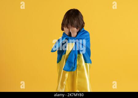 Unhappy little boy wrapped in Ukrainian flag crying, hiding face in hands, suffering from fear and pain, free space Stock Photo
