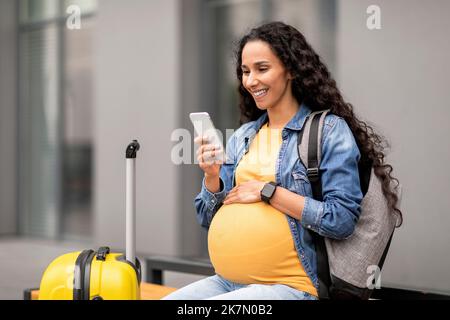 Cheerful young pregnant woman travelling alone, using smartphone at airport Stock Photo