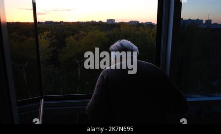 Senior caucasian woman grandmother standing near window looking outdoor on green forest in sunset isolation or quarantine and shaking hand for greetin Stock Photo