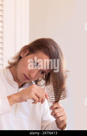 Vertical view of young long haired man taking care and brushing his hair with anti fall hair brush Stock Photo