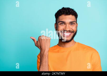 Photo of pretty charming guy wear orange t-shirt pointing thumb empty space smiling isolated teal color background Stock Photo