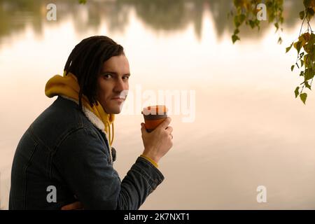 Young handsome man with dreadlocks in warm clothes drinking coffee near lake in golden autumn Stock Photo