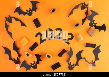 Composition on Halloween from bats cut from paper and various candies, on an orange background. In the center inscription 2022 Stock Photo