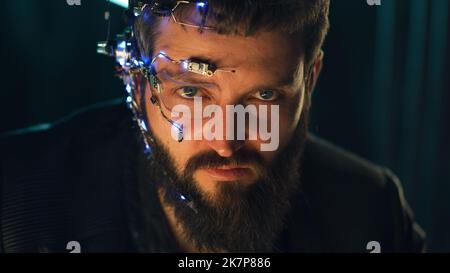 Brunette guy with dark beard wearing futuristic headset with one-eyed glasses, mic and ear piece looks at the camera with intense facial expressions. Neon lights in the background. Cyberpunk. Stock Photo