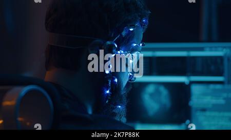 Back view of a brunette guy wearing futuristic headset with LED lights attached programs and develops code using multiple computer screen. Cyperpunk style. Sci-fi background. Neon lights. Stock Photo