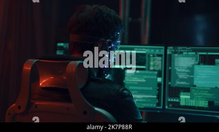 Back view of a brunette guy wearing futuristic headset with LED lights attached programs and develops code using multiple computer screen. Cyperpunk style. Sci-fi background. Neon lights. Stock Photo