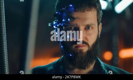 Brunette guy with dark beard wearing futuristic headset with one-eyed glasses, mic and ear piece looks at the camera with intense facial expressions. Neon lights in the background. Cyberpunk. Stock Photo