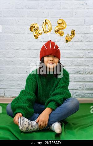Funny Asian girl in a green sweater sits on the floor with golden balloons with the number 2023 in a red cap. A girl sits cross-legged on the floor wi Stock Photo