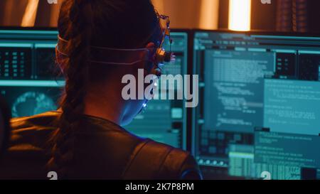 Girl works with multiple computer screen. Wearing a headset with LED lights programming on computer. Coding. Braided haired girl in cyberpunk style. Stock Photo