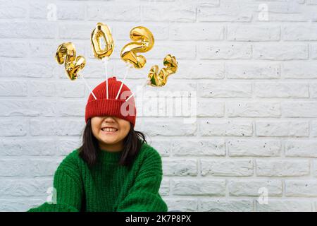 Happy Asian baby girl in a green sweater close-up with golden balloons with numbers 2023 in hat. Copy space, background of a white brick wall, the eye Stock Photo