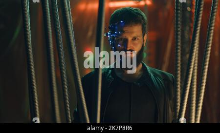 Young brunette guy in Cyberpunk style stands behind the black rods hanging. Wearing a futuristic headset with eyepiece and microphone. Neon lights background. Science fiction. Stock Photo