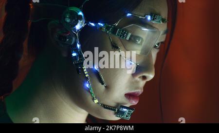 Focused asian girl in cyberpunk attire works on the computer. Wearing one-eyed glasses with white LED lights and a microphone. Neon lights background. science fiction, cybernetics concept. Stock Photo