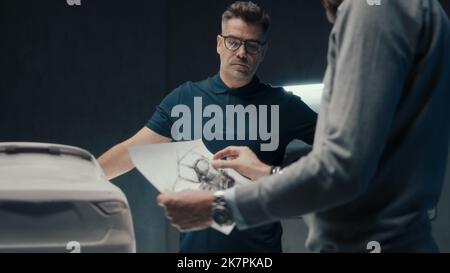 Experienced car designers and developers discuss the design of the car while standing infront of the sculpture of prototype car. Using the pencil sketches in a high tech car manufacturing company. Stock Photo