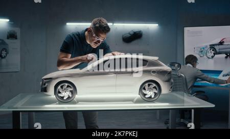 Senior automotive engineers work in high tech car manufacturing company. One of them works on the sculpture of the prototype car while the other one uses a digital touch screen table to draw sketches. Stock Photo