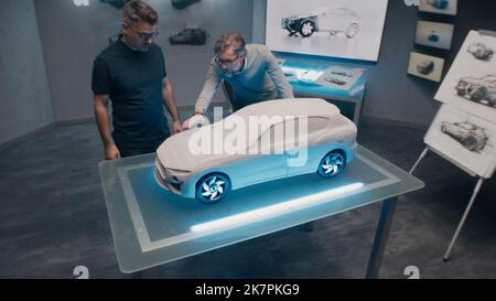 Senior automotive developers make the design corrections looking at the sculpture of the prototype car model on a glass table. High tech office with LED and car sketches. Stock Photo