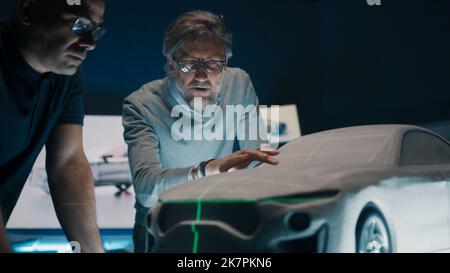Senior automotive engineers discuss the car design infront of a sculpture prototype car model using a green laser light for perfection. Model car placed on a glass table in a high tech laboratory. Stock Photo