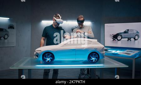 Automotive professional engineers with VR headset and joysticks talk about vehicle production while standing infront of a prototypecar model in a high tech office. Car design analysis and improvement. Stock Photo