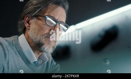 A middle aged senior automotive design engineer with black framed glasses draws sketch of the prototype model of a car. Automotive industry. Modern studio. Stock Photo