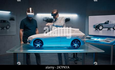 Automotive professional engineers with VR headset and joysticks talk about vehicle production while standing infront of a prototypecar model in a high tech office. Car design analysis and improvement. Stock Photo