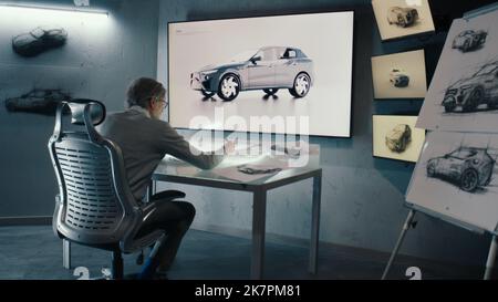 Senior automotive graphic designer works with digital display graphics touch sreen table. Looks at the rotating 3d image of the car in a wall mounted LED. High tech innovative laboratory. Stock Photo