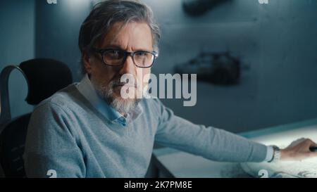 Closeup face of a senior automotive graphic designer with black framed glasses and beard stops work and looks at the camera. Sketches of prototype model car in a high tech innovative laboratory. Stock Photo