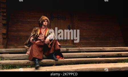 Pensive young ukrainian cossack dressed traditional clothing holding saber in his hand sitting on the steps and looking into the distance Stock Photo