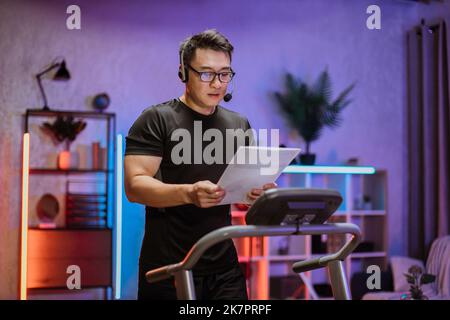 Side view of asian man in glasses and sports clothes using headset to work with papers while doing cardio training on treadmill at night at home or gym. Concept of sport, health care, business. Stock Photo