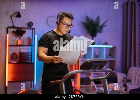 Portrait of young active asian businessman in grey sportswear and headset training at home working with papers, doing cardio exercise on treadmill at evening time. Concept of sport, business. Stock Photo