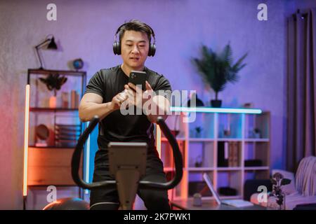 Young asian man with headphones doing exercises on stationary bicycle at home or fitness center. Young sporty male in gym listen music from smartphone. Man doing cardio exercises. Stock Photo