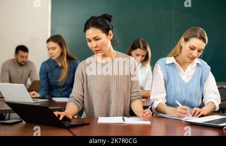 Students writing in notebook and typing on laptop during class in classroom Stock Photo