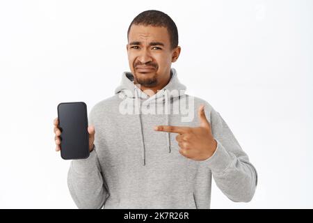 Disappointed guy points at his mobile phone screen, shows telephone and grimaces with dislike, looks upset, stands over white background Stock Photo