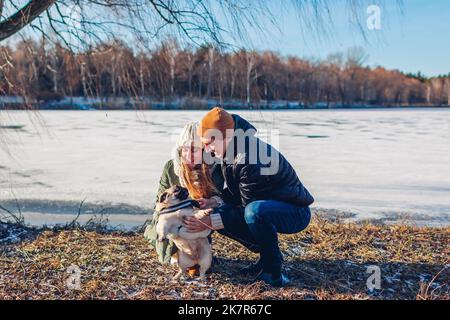 Young couple walking pug dog in snowy winter park. Man and woman playing with pet outdoors having fun. Activities with animals Stock Photo