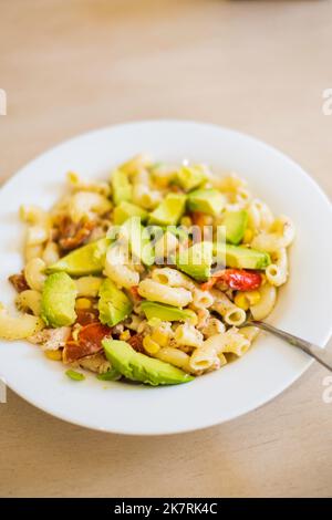 A cold pasta salad consisting of large elbow macaroni, slice avocados, toasted red bell peppers and corn in a white dish sat on a wood table. USA. Stock Photo