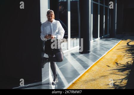 A plus-size Hispanic businesswoman leaning against the black wall of an office building exterior with a strict glass facade and using her smartphone, Stock Photo